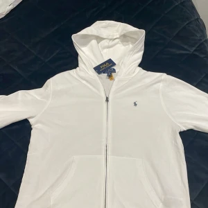 Vit hoodie från Polo Ralph Lauren| XL 18-20 år - Säljer en stilren vit hoodie från Polo Ralph Lauren. Den har en dragkedja framtill och en liten broderad logotyp på bröstet. Perfekt för en avslappnad look. Kommer i originalförpackning. Lapparna sitter kvar