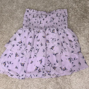 Lila blommig kjol - Söt lila kjol med blommigt mönster och volanger. Perfekt för en somrig look med sin medel längd och elastiska midja. Kjolen har ett romantiskt och lekfullt uttryck.