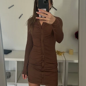 Brun långärmad klänning - Snygg brun klänning med långa ärmar och knappar framtill. Den har en åtsittande passform med rynkdetaljer som ger en smickrande siluett. Perfekt för en stilren look.