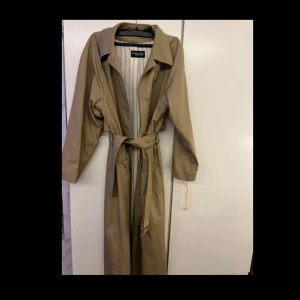 Beige trenchcoat  - Snygg beige trenchcoat med knytbälte i midjan och klassisk krage. Jackan har långa ärmar och en stilren design med randigt foder. Perfekt för höst och vår!