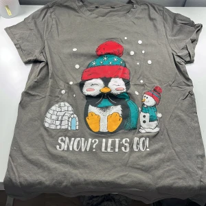 Grå t-shirt med pingvinmotiv - Säljer en grå t-shirt med ett gulligt pingvinmotiv. T-shirten har korta ärmar och texten 'SNOW? LET'S GO!' under motivet. Perfekt för vintern med sitt charmiga tryck av en pingvin i mössa och halsduk. Är i storlek xs och är från New yorker. Passar även barn 
