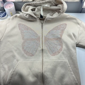 Beige hoodie med fjärilsdetalj - Säljer en beige hoodie med dragkedja och en stor fjärilsdetalj i glittrande stenar på framsidan. Hoodien har en mysig huva och praktiska fickor framtill. Storlek xs (liten i storleken och kort i ärmarna.pris kan diskuteras🦋✨