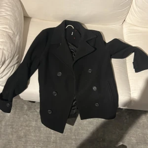 Svart skepparkavaj från Pier One - Snygg svart skepparkavaj från Pier One med dubbelknäppning och klassisk krage. Jackan har långa ärmar och är tillverkad i polyester. Perfekt för en stilren look.