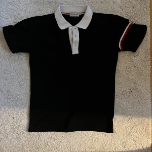 Svart pikétröja från Moncler S/M - Säljer en stilren svart pikétröja från Moncler med vit krage och knappar. Tröjan har korta ärmar och en snygg detalj med Moncler-logga på ärmen. Perfekt för en avslappnad men ändå elegant look. Unik. Gammal model så NFC fungerar ej. Vid frågor skriv vi kan ta ner priset!!! Passar som en tajt M närmare S!!!
