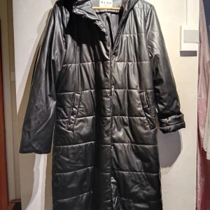 Svart lång dunjacka från NA-KD - Säljer en svart lång dunjacka från NA-KD med huva och dragkedja. Jackan har långa ärmar och quiltad design, perfekt för kyliga dagar. Den har praktiska fickor och en stilren look. Passar utmärkt för vintern!