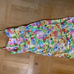 Färgglad blommig klänning - Säljer en färgglad klänning med blommigt mönster i olika nyanser av rosa, grön, gul och blå. Klänningen har smala axelband och är perfekt för sommarens varma dagar. Den är tillverkad i ett lätt och luftigt material.
