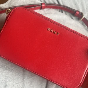 Röd axelväska från DKNY - Snygg röd axelväska från DKNY i skinn med guldfärgade detaljer. Väskan har en praktisk dragkedja och en justerbar axelrem. Perfekt för att ge en färgklick till din outfit!