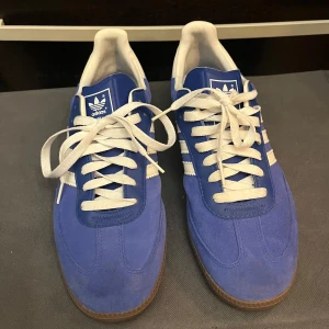 Blå Adidas Samba sneakers - Snygga blå Adidas Samba sneakers med vita detaljer och snörning. Skorna har en klassisk design med tre vita ränder på sidan och Adidas-logga på tungan och hälen. Perfekta för en sportig look. Nya utan kartong 