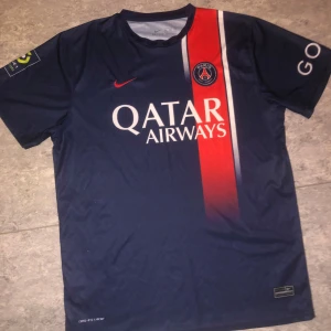 Blå fotbollströja från Nike - Säljer en blå PSG-fotbollströja från Nike med röd och vit rand på framsidan. Tröjan har korta ärmar och sponsorloggor på bröstet och ärmarna. Perfekt för fotbollsfans! Står Xl men är själv ett L och den passar perfekt