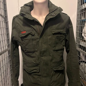 Grön fieldjacka från Superdry - Säljer en grön fieldjacket från Superdry. Jackan har flera praktiska fickor och en dragkedja framtill. Den har en cool patch på ärmen och är perfekt för höst och vår. Storlek large.