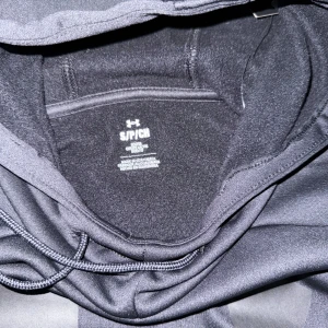 Svart hoodie från Under Armour - Säljer en stilren svart hoodie från Under Armour med stor logga på bröstet. Tröjan har en praktisk känguruficka och justerbar huva med dragsko. Perfekt för både träning och vardag. 🖤