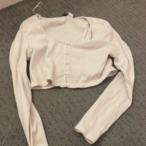 Vit ribbad croppad kofta - Säljer en stilren vit ribbad kofta med knappar framtill. Den är croppad och har långa ärmar, perfekt för lager-på-lager-stil. Passar både till jeans och kjol för en trendig look.