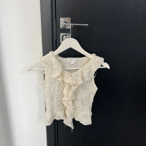 Beige volangblus - Charmig beige blus med volangdetaljer framtill. Blusen är ärmlös och har en lätt textur i tyget som ger en elegant touch. Perfekt för varmare dagar eller som ett lager under en kavaj.