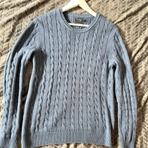 Blå stickad tröja från East West - Säljer en stilren blå stickad tröja från East West. Tröjan har ett klassiskt kabelstickat mönster och rund halsringning. Perfekt för kyligare dagar och passar bra till både jeans och chinos. Passar även M.  Tröjan är mörkare än på bilderna.