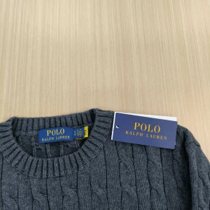 Grå kabelstickad tröja från Polo Ralph Lauren - Säljer en stilren grå kabelstickad tröja från Polo Ralph Lauren. Tröjan har en klassisk rund hals och är prydd med det ikoniska lila logotypbroderiet på bröstet. Perfekt för en elegant och avslappnad look. Den är även helt ny och ingen skada, det är storlek s men den passar även m
