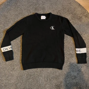 Svart tröja från Calvin Klein Jeans - Snygg svart tröja från Calvin Klein  med vita detaljer på ärmarna där märkesnamnet står. Tröjan har långa ärmar och en rund halsringning. Perfekt för en stilren look.