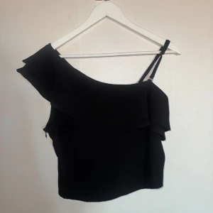 Svart oneshoulder topp från Rut&Circle - Snygg svart oneshoulder topp från Rut&Circle med volangdetalj. Perfekt för en stilren look. Toppen har en smal axelrem och är croppad för en modern stil.