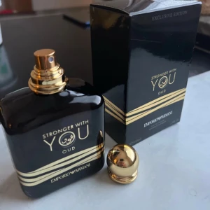 Stronger With You Oud från Emporio Armani - Exklusiv parfym 'Stronger With You Oud' från Emporio Armani. Flaskan är elegant i svart med gyllene detaljer och en matchande gyllene kork. Förpackningen är också svart med gyllene inslag, vilket ger en lyxig känsla. Perfekt för den som söker en sofistikerad doftupplevelse.