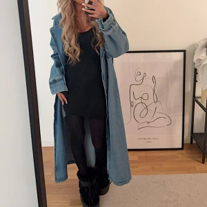 Lång jeanskappa - Säljer en lång och stilren jeanskappa i ljusblå denim. Kappan har en avslappnad passform med knappar framtill och långa ärmar. Perfekt för en trendig look året runt.