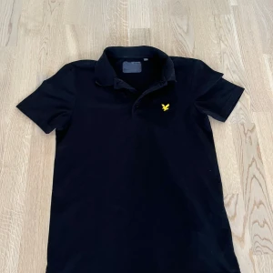 Svart pikétröja från lyle & scott  - Snygg svart pikétröja från lyle & scott som har korta ärmar och en krage med knappar.