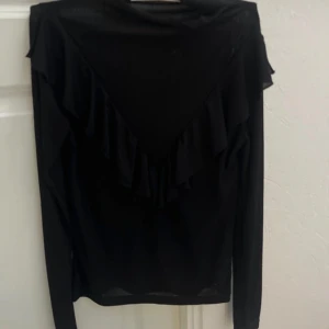 Svart långärmad topp med volang - Snygg svart långärmad topp med volangdetaljer. Perfekt för en stilren look. Toppen har en elegant design med volanger som ger en extra touch.