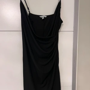 Svart klänning med drapering - Elegant svart klänning med smala axelband och vacker drapering framtill. Klänningen har en omlott-design som ger en smickrande passform.