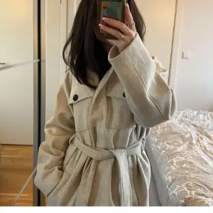 Säljer en superfin beige kappa med bälte i midjan. Den har stora knappar och två bröstfickor som ger en snygg look. Perfekt för höst och vår när det är lite kyligt ute. Kappan är långärmad och har en klassisk krage.