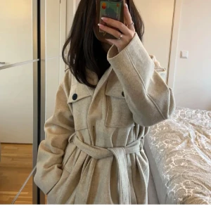 Beige kappa med bälte - Säljer en superfin beige kappa med bälte i midjan. Den har stora knappar och två bröstfickor som ger en snygg look. Perfekt för höst och vår när det är lite kyligt ute. Kappan är långärmad och har en klassisk krage.