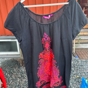 Svart klänning med rosa mönster från Indiska - Säljer en snygg svart tunika från Indiska med ett coolt rosa mönster framtill. Klänningen har korta ärmar och en bekväm passform, perfekt för både vardag och fest. Den är gjord i ett lätt och luftigt material, vilket gör den idealisk för sommardagar.