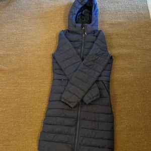 Mörkblå lång pufferjacka från CRW - Säljer en snygg och varm mörkblå pufferjacka från CRW. Jackan har en lång modell med huva och dragkedja framtill. Den är perfekt för kalla dagar och har praktiska fickor med dragkedja. Passar perfekt för både vardag och ridning.