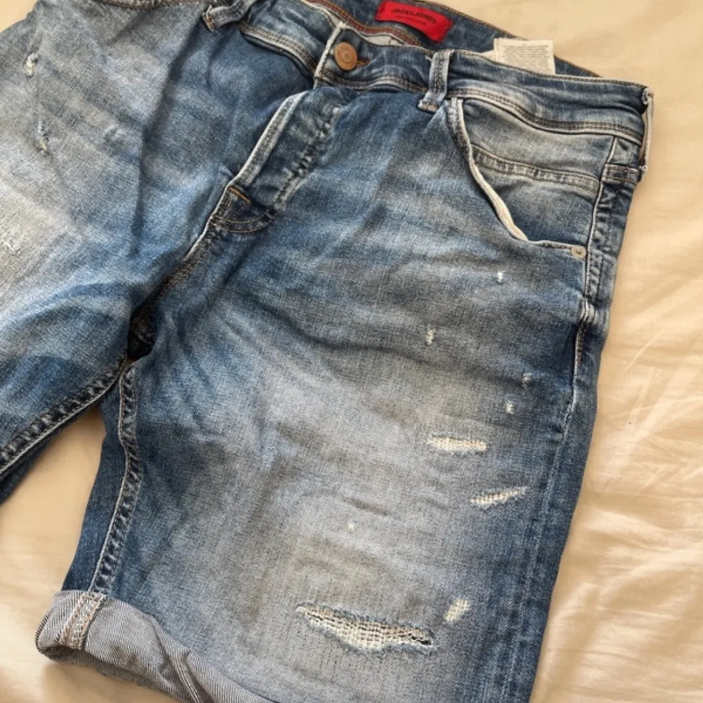 Säljer ett par snygga blå jeansshorts från Jack & Jones. De har en cool slitning och är i en klassisk femficksmodell. Perfekta för sommaren med en avslappnad stil. Shortsen har en knapp och dragkedja framtill.. Shorts.