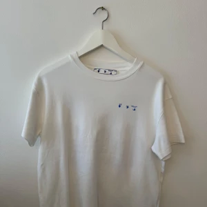 Vit t-shirt från Off-White med blommigt tryck - Säljer en snygg vit t-shirt från Off-White i storlek medium. Den har ett coolt blommigt tryck på baksidan med blå detaljer. Perfekt för sommaren och att styla med jeans eller shorts. T-shirten är i bomull och har en normal passform.