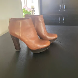 Bruna stövletter med klack och dragkedja - Säljer ett par snygga bruna stövletter från Michael Kors i nyskick. De är i skinn och har en hög klack som ger en elegant look. Dragkedjan i guld på baksidan gör dem lätta att ta på och av. Perfekta för höst och vinter!