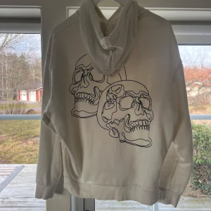 Vit hoodie med dödskallemotiv - Säljer en stilren vit hoodie med ett coolt dödskallemotiv på ryggen. Tröjan har en klassisk passform med huva och dragsko samt en praktisk magficka. Perfekt för en avslappnad stil. Storlek M men känns mer som S