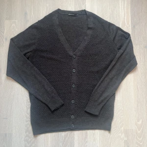 Mörkgrå kofta från DeFacto Studio - Säljer en stilren mörkgrå kofta från DeFacto Studio. Koftan har en v-ringad design med knappar framtill och ett subtilt ribbat mönster. Perfekt för kyligare dagar.