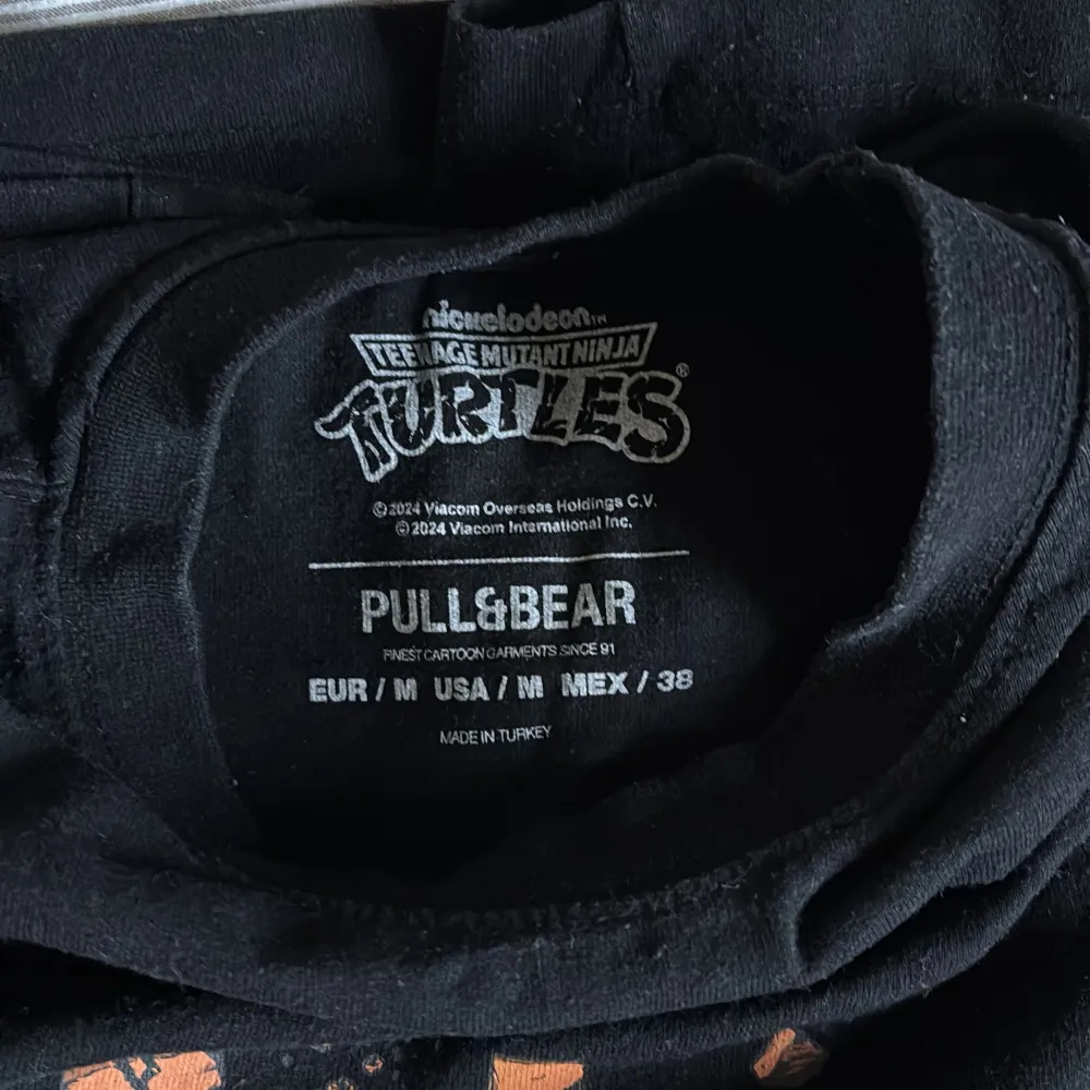 Cool svart oversized t-shirt med Teenage Mutant Ninja Turtles-tryck från Pull&Bear. T-shirten har ett färgglatt motiv av de fyra sköldpaddorna på baksidan och en mindre logga på framsidan. Perfekt för fans av den klassiska serien!. T-paidat.