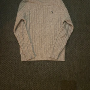 Grå stickad tröja från Polo Ralph Lauren - Säljer en stilren grå stickad tröja från Polo Ralph Lauren med kabelstickat mönster och broderad logotyp på bröstet. Tröjan har långa ärmar och en rund halsringning. Perfekt för kyligare dagar. Sitter perfekt runt midjan och armarna jävligt skönt material då det är kasmhier tröja skulle säga att den passar alla från S till M och ja den är äkta