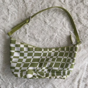 Grön och vit axelväska från Monki - Snygg axelväska från Monki i grönt och vitt med rutigt mönster. Väskan har en justerbar axelrem och stängs med dragkedja. Perfekt för att ge en färgklick till din outfit!