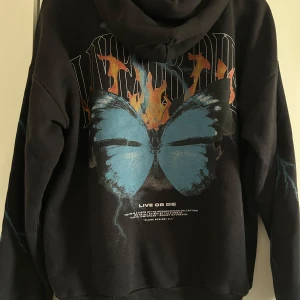 Svart hoodie med tryck - Säljer en svart hoodie med ett stort tryck av en blå fjäril och eldflammor på baksidan. Framsidan har texten 'ALONE against ALL'. Hoodien har en stor ficka framtill och ribbade muddar vid ärmslut och nederkant.