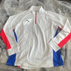 Vit långärmad tröja från Puma - Säljer en vit långärmad tröja från Puma med dragkedja vid halsen. Tröjan har röda och blå detaljer på ärmarna och sidorna. Perfekt för sportiga aktiviteter eller en avslappnad stil.