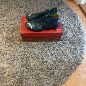 Blå fotbollsskor från Puma. Elite model - Snygga blå fotbollsskor från Puma med vit logga och snörning. Skorna har en modern design med detaljer i svart och grönt. Perfekta för fotbollsplanen med en tight passform som ger bra kontroll. Nypris 2599 box ingår 