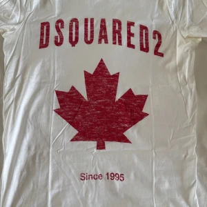 Vit t-shirt från Dsquared2 med rött lönnlöv - Säljer en vit t-shirt från Dsquared2 med ett stort rött lönnlöv och texten 'Since 1995' tryckt på framsidan. Perfekt för en avslappnad stil med en touch av kanadensisk inspiration.