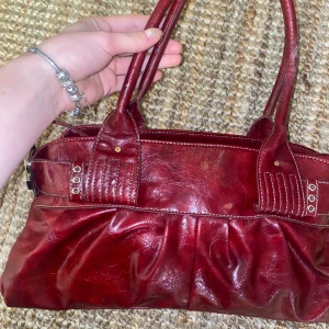 Röd skinnhandväska med detaljer - Snygg röd handväska i skinn med dekorativa spännen och nitar. Väskan har dubbla handtag och en stilren design som passar perfekt för att lyfta din outfit. Perfekt för dig som vill ha en praktisk men ändå trendig accessoar.