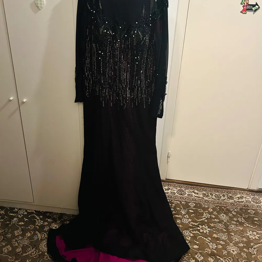 Elegant svart och lila långklänning med glittrande paljetter och detaljerad brodering. Klänningen har långa ärmar och en vacker fallande kjol med en hint av lila underton. Perfekt för en glamorös kväll.. Mekot.