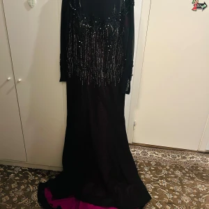 Svart långklänning med paljetter - Elegant svart och lila långklänning med glittrande paljetter och detaljerad brodering. Klänningen har långa ärmar och en vacker fallande kjol med en hint av lila underton. Perfekt för en glamorös kväll.