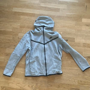 Nike tech  - Säljer en stilren grå hoodie från Nike med dragkedja och huva. Tröjan har långa ärmar och svarta detaljer vid dragkedjan och ärmsluten. Perfekt för en avslappnad look eller träning.