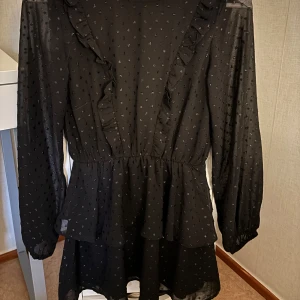 Svart klänning med volangdetaljer  - Elegant svart klänning med långa ärmar och volangdetaljer längs framsidan. Klänningen har ett diskret prickigt mönster och en luftig passform som ger en fin siluett. Perfekt för en stilfull look. Den är helt ny klänning helt fräs.