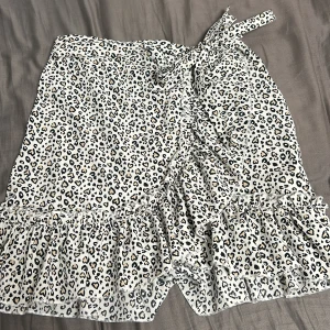 Leopardmönstrad omlottkjol - Snygg leopardmönstrad omlottkjol med volangdetaljer och knytband i midjan. Kjolen har en dragkedja på sidan för enkel på- och avtagning. Perfekt för att ge en trendig touch till din outfit. Köptes utomlands så jag vet inte märket. Säljs på grund av liten användning.