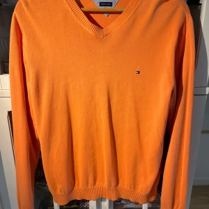 Orange tröja från Tommy Hilfiger - Säljer en snygg orange tröja från Tommy Hilfiger i premium bomull. Tröjan har en v-ringad design och långa ärmar, perfekt för en stilren look. Den har en liten logotyp på bröstet och ribbade kanter för extra komfort. Finns små fläckar som ni ser på bilderna