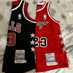 Basketlinnen från Mitchell & Ness - Två basketlinnen från Mitchell & Ness med nummer 23. Ena linnet är svart med vita och röda detaljer, det andra är rött med vita och svarta detaljer. Båda har stjärnor på sidan och NBA-loggan på bröstet. Perfekt för basketfans!Pris kan diskuteras!!